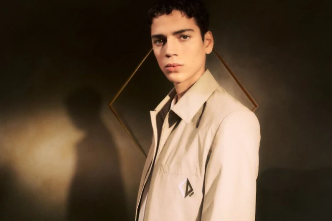 Portrait of a young adult male in a beige jacket, posing in front of a framed diamond shape, with a dark greenish-brown background.