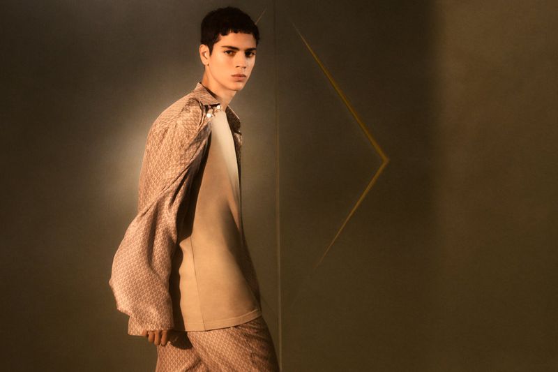 Young man poses in a patterned shirt, light jacket, and matching pants in a studio with earth-tone colors and geometric light.