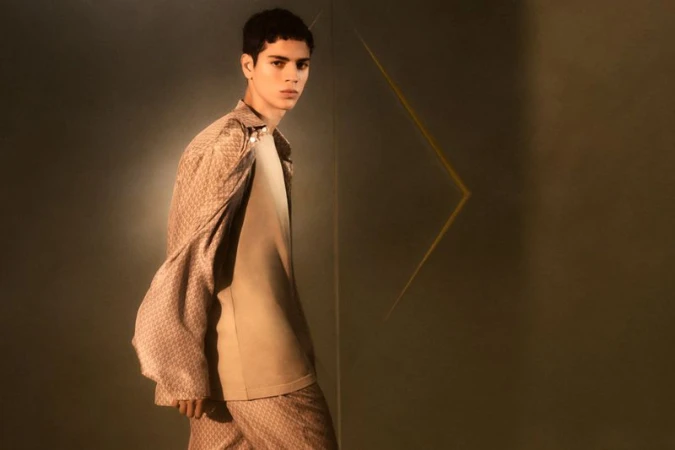 Young man poses in a patterned shirt, light jacket, and matching pants in a studio with earth-tone colors and geometric light.