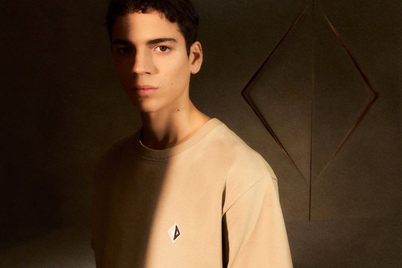 Portrait of a young adult male in a beige t-shirt, looking at the camera with a neutral expression, against a dark background with geometric details.