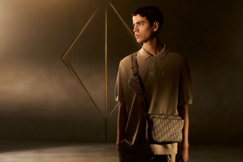 Young adult male poses indoors with a tan polo shirt and designer crossbody bag in a diffused, warm light setting, creating a fashion-forward look.