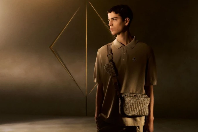 Young adult male poses indoors with a tan polo shirt and designer crossbody bag in a diffused, warm light setting, creating a fashion-forward look.
