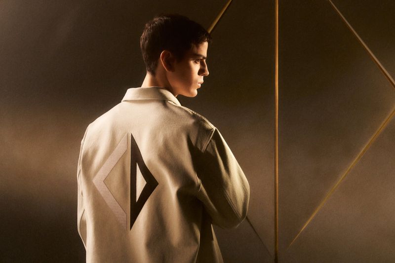 Side profile of a young adult male with short hair wearing a light jacket with a dark geometric design on the back in a studio setting.