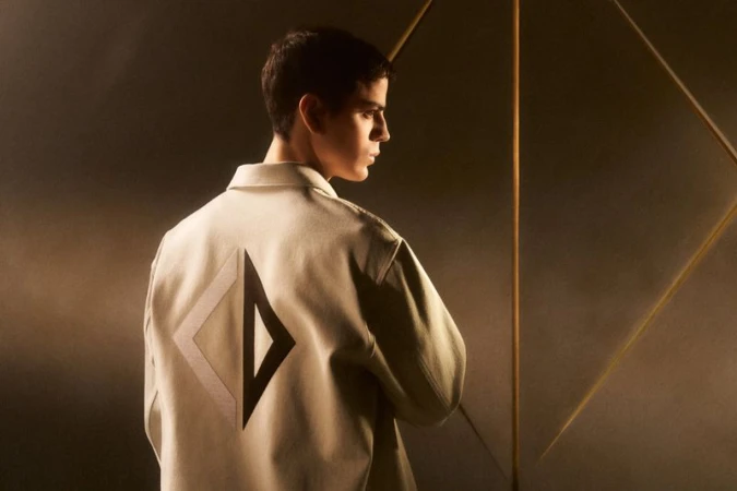 Side profile of a young adult male with short hair wearing a light jacket with a dark geometric design on the back in a studio setting.
