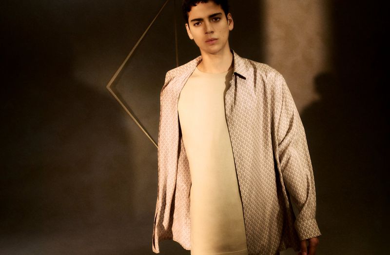 Portrait of a young man in casual beige shirt and patterned jacket, gazing at the camera against a neutral brown background.