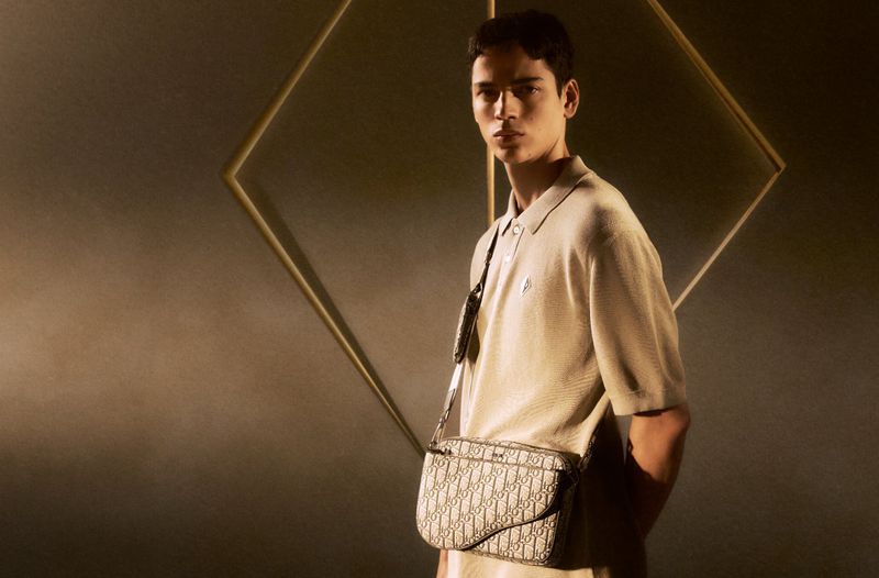 A stylish young adult man is posing in a light brown polo shirt with a branded messenger bag against a dark backdrop with a geometric frame.