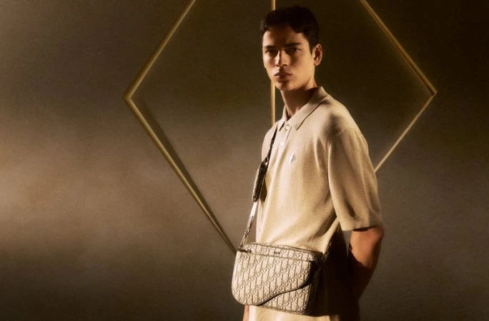 A stylish young adult man is posing in a light brown polo shirt with a branded messenger bag against a dark backdrop with a geometric frame.
