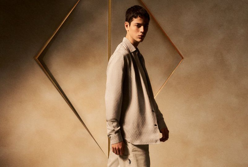 A young adult male in casual fashion poses in front of a geometric gold frame against a textured wall. The setting is a neutral-toned photo studio.