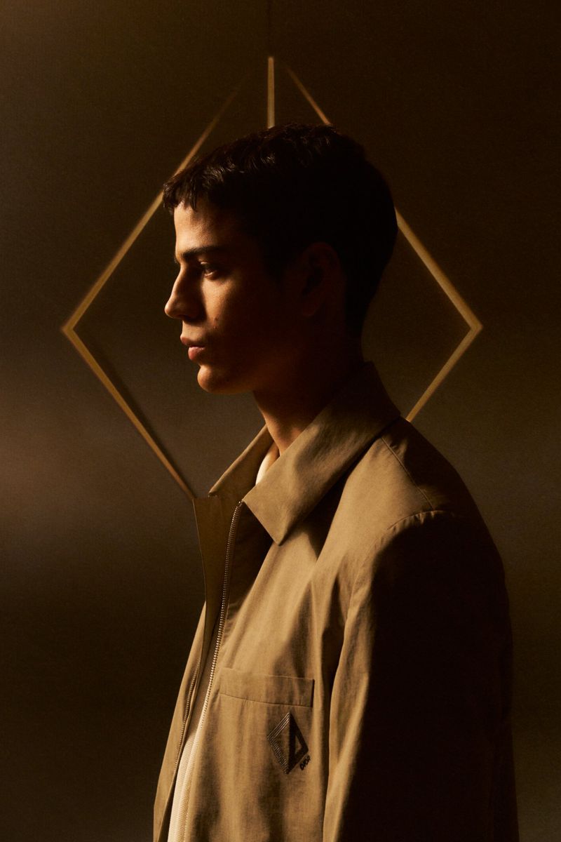 Profile of an adult male in a tan jacket in a studio, featuring a geometric background. Soft, warm lighting and shallow depth of field.