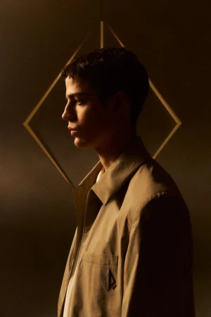 Profile of an adult male in a tan jacket in a studio, featuring a geometric background. Soft, warm lighting and shallow depth of field.