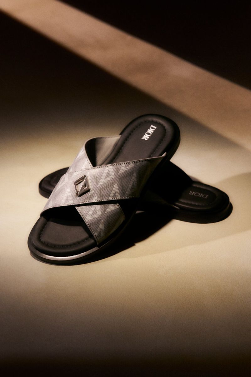 Studio shot of Dior men's slide sandals featuring criss-cross patterned straps and signature Dior logo on a neutral background.