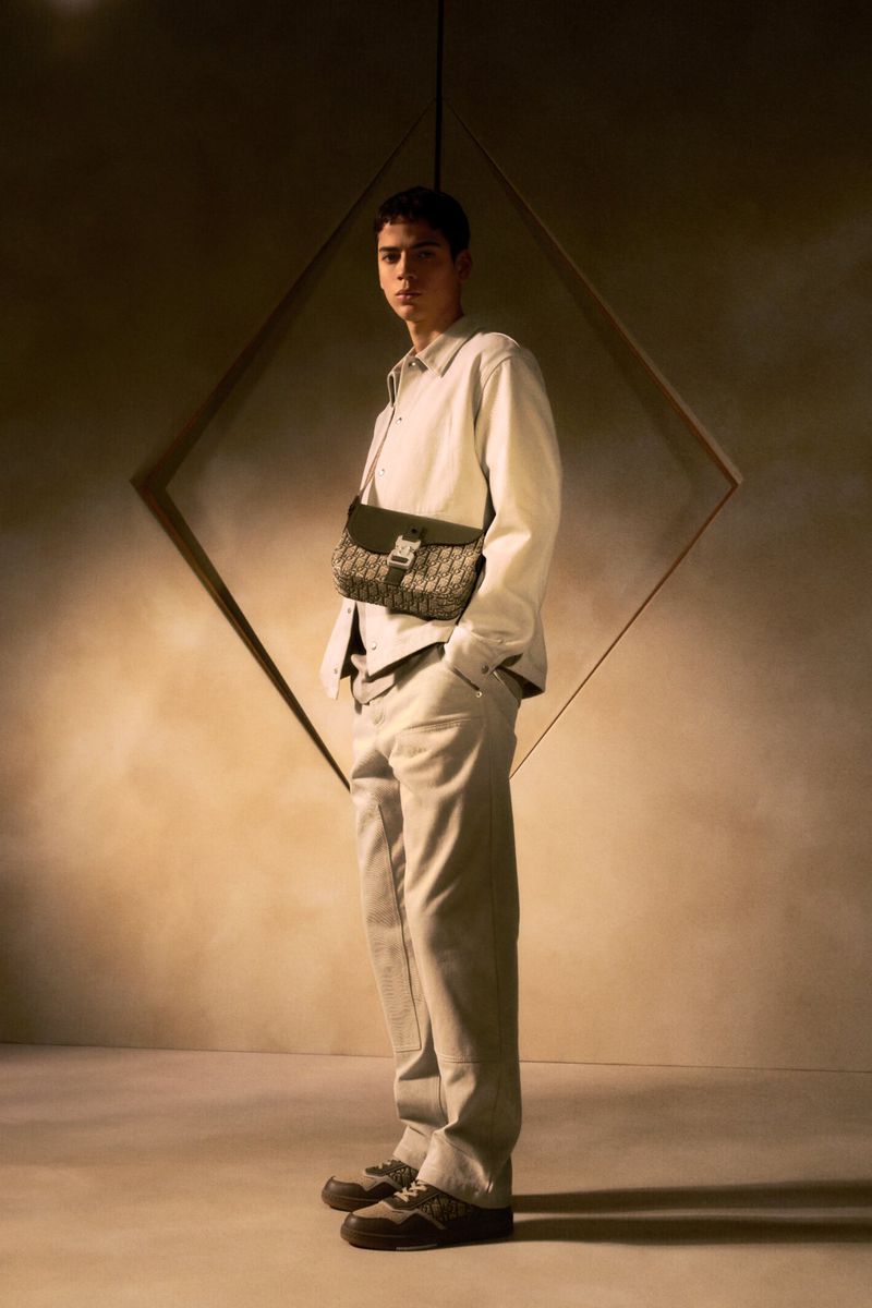 Man in cream outfit stands in front of a geometric wall design in a fashion shot, wearing a brown patterned bag and matching sneakers.