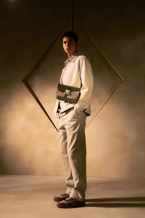 Man in cream outfit stands in front of a geometric wall design in a fashion shot, wearing a brown patterned bag and matching sneakers.