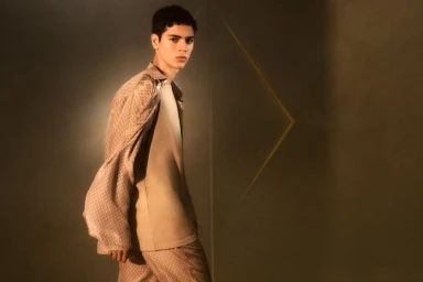 Model wearing a patterned patterned brown shirt and trousers against a dark wall with geometric gold line detailing.