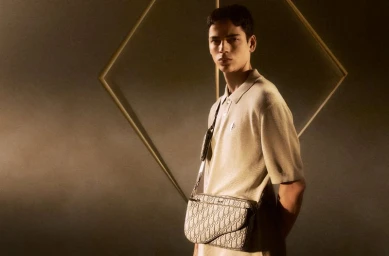 Model wearing a beige polo shirt and carrying a patterned crossbody bag against a textured warm-lit background.