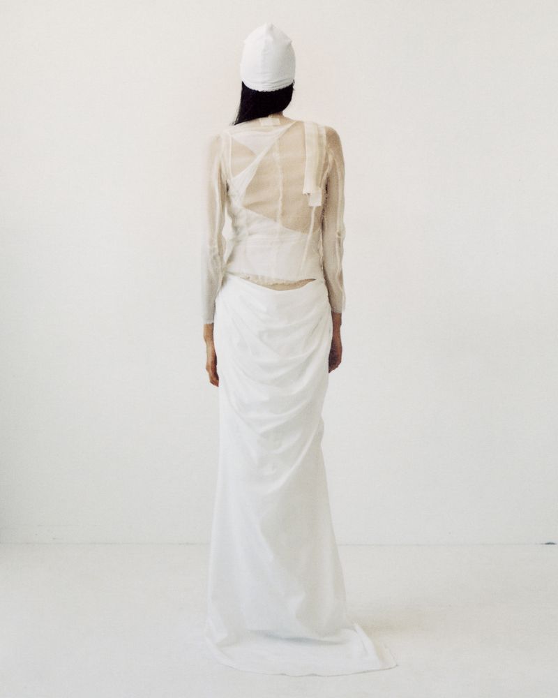 Back view of an adult female wearing a white gown and cap standing in a white photo studio setting.