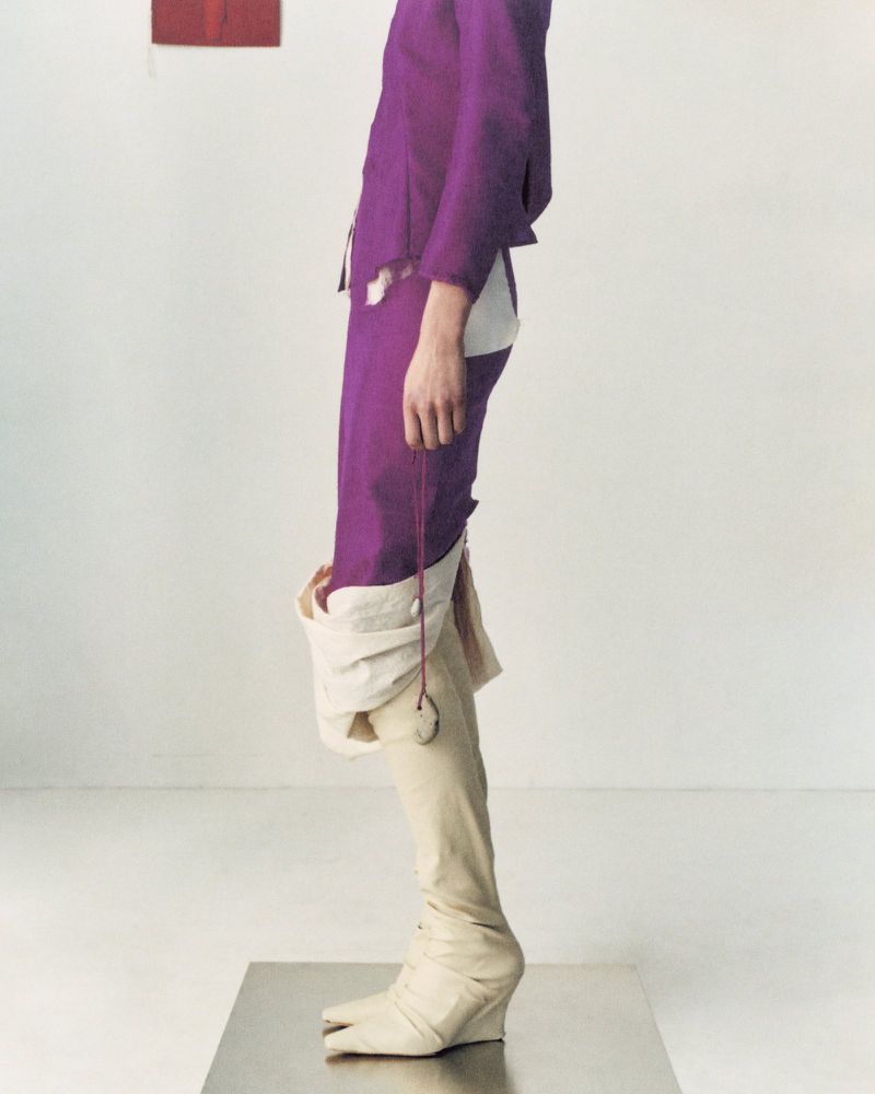 Fashionable individual wearing a purple suit with unique split-toe boots strikes a pose in a stark white photo studio setting.