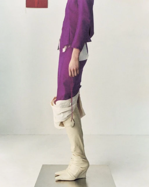 Fashionable individual wearing a purple suit with unique split-toe boots strikes a pose in a stark white photo studio setting.
