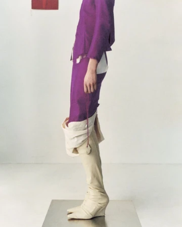 Fashionable individual wearing a purple suit with unique split-toe boots strikes a pose in a stark white photo studio setting.