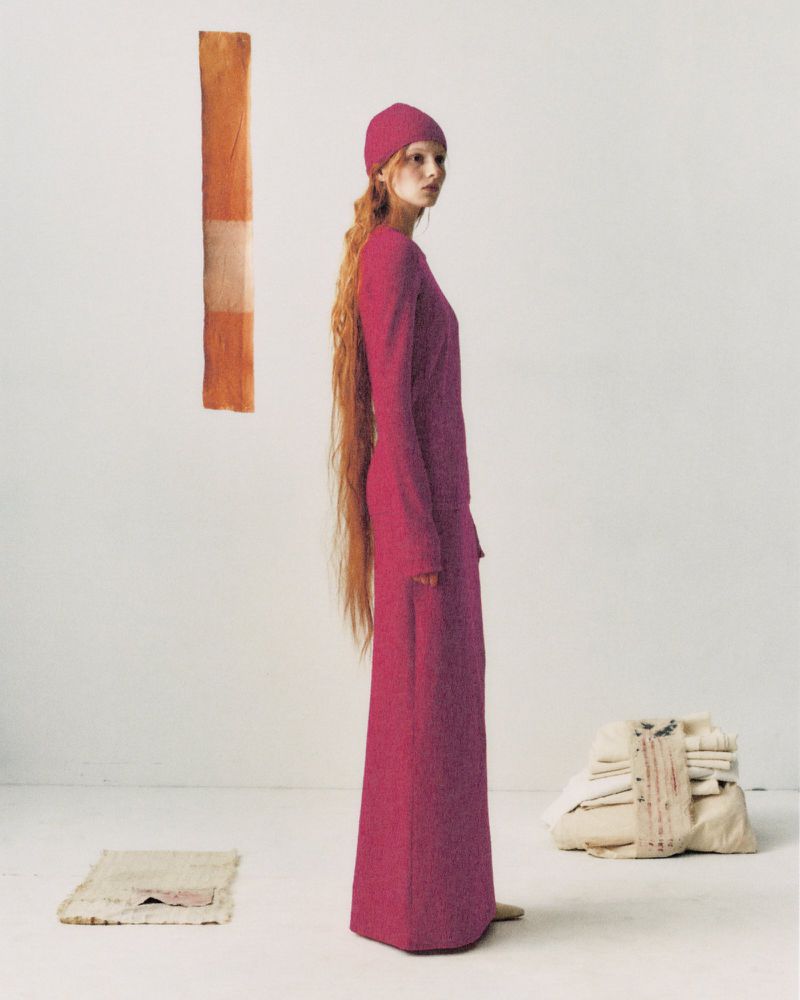 A slender woman with long red hair poses in a pink dress and beanie in a minimal studio setting with textiles and neutral tones.