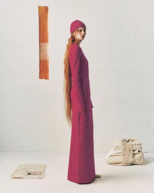 A slender woman with long red hair poses in a pink dress and beanie in a minimal studio setting with textiles and neutral tones.