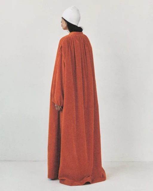 Back view of a fashion model in an orange oversized robe and white hat, standing against a white backdrop in a studio setting.