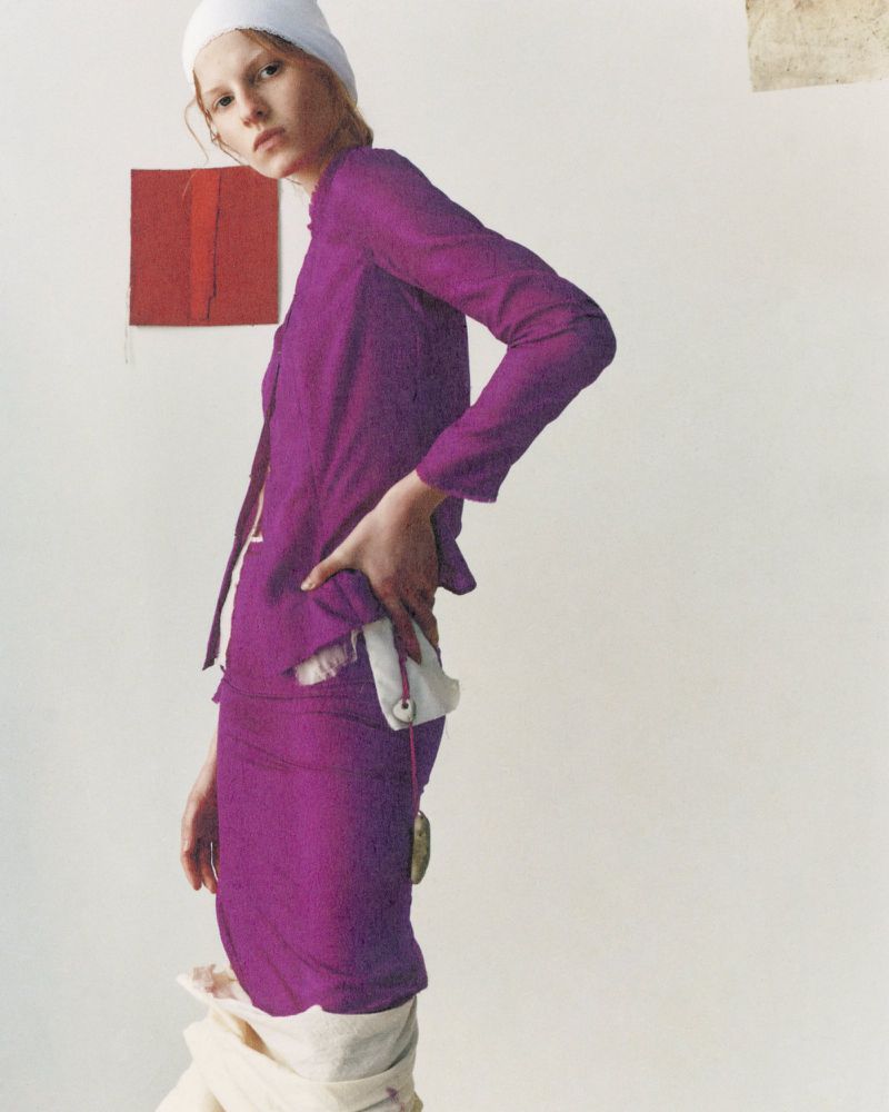 Woman posing in a purple suit with a white headscarf against a plain backdrop with color swatches, fashion editorial shot.