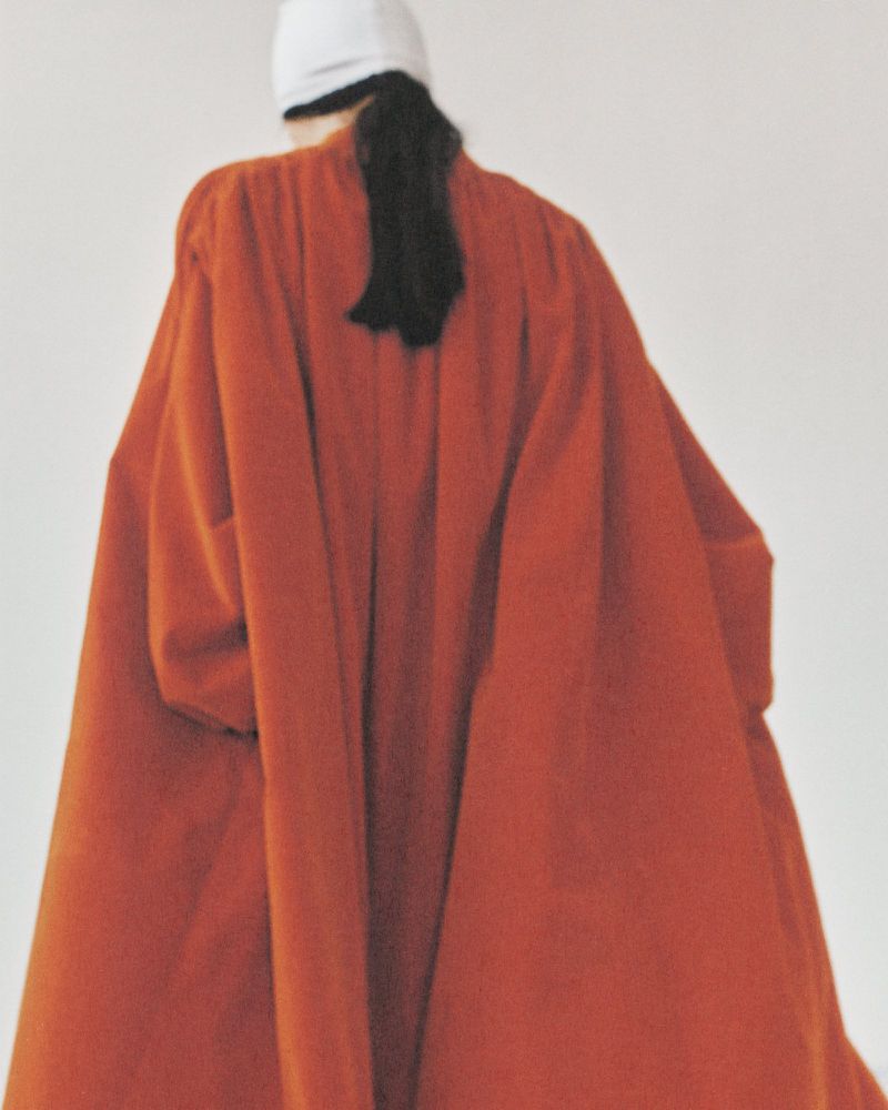 Back view of a person wearing a rust-colored oversize coat and a white baseball cap, posing against a plain white background.