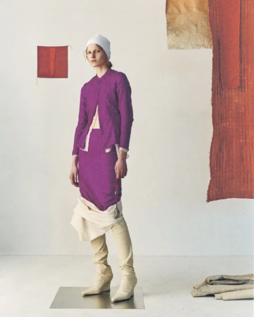 A woman in a purple skirt suit and white beanie stands in a studio with textiles hanging in the background, full-length fashion portrait.