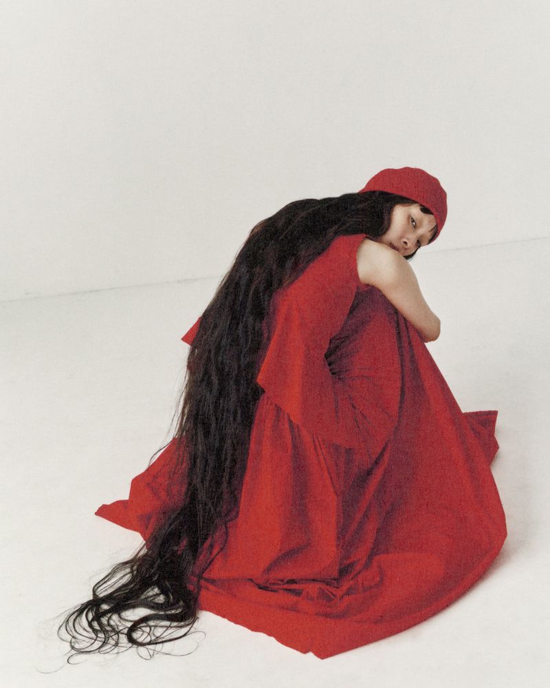A single woman crouches in a photo studio wearing a red dress and matching cap, with very long dark hair flowing onto the white floor.
