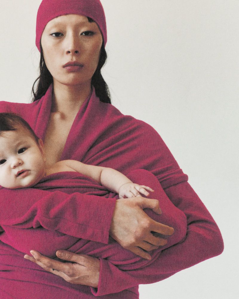 An Asian woman cradles a baby in a magenta wrap in a studio shot with a simple background, creating a soft, maternal ambiance.