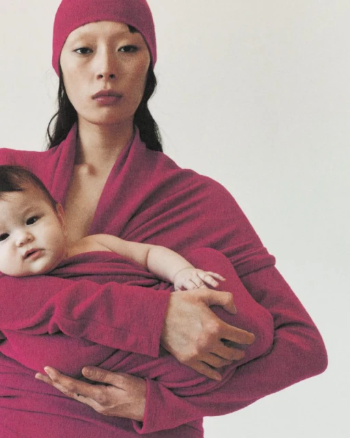 An Asian woman cradles a baby in a magenta wrap in a studio shot with a simple background, creating a soft, maternal ambiance.
