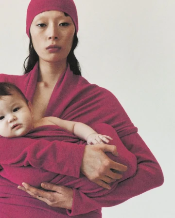 An Asian woman cradles a baby in a magenta wrap in a studio shot with a simple background, creating a soft, maternal ambiance.