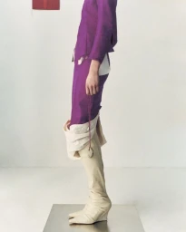 Cropped studio view of a model in a vibrant purple suit and draped cream knee-high boots standing atop a metallic platform.
