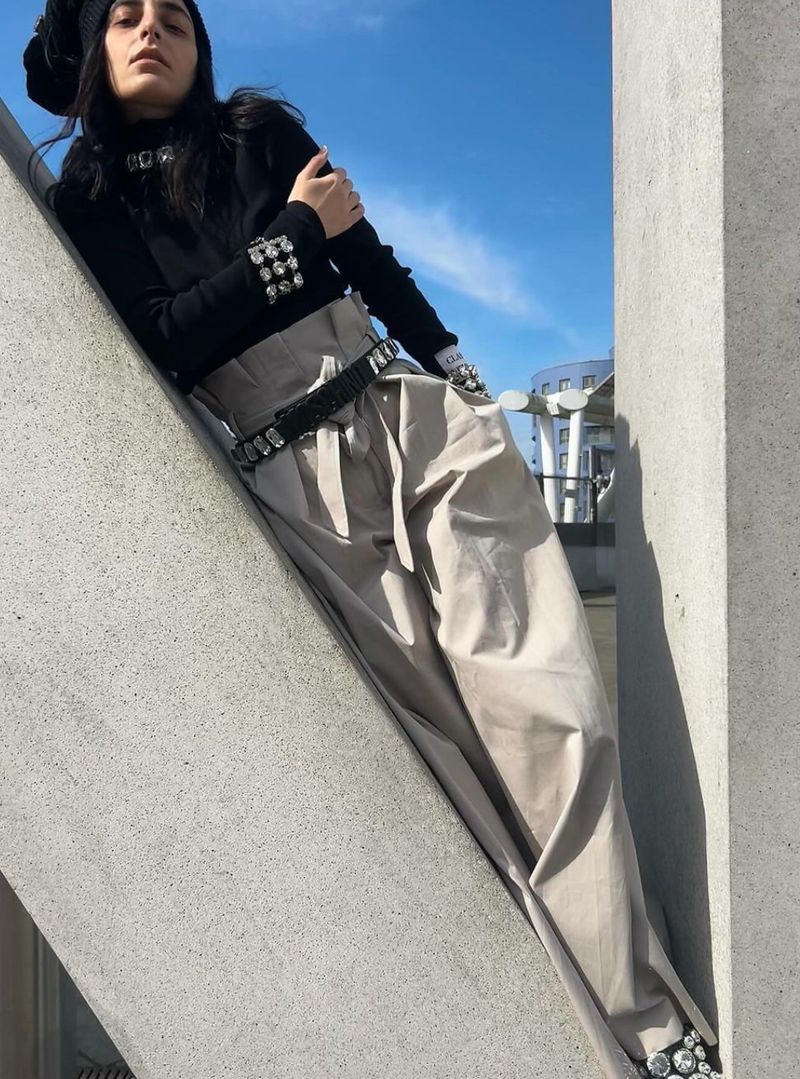A fashionable young woman strikes a pose outside, wearing beige pleated trousers, a black knit top, and studded accessories on a sunny day.