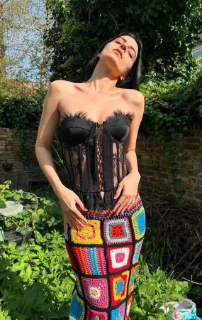 Adult female poses in a black corset top and a colorful crochet skirt in an outdoor garden setting, under natural sunlight.