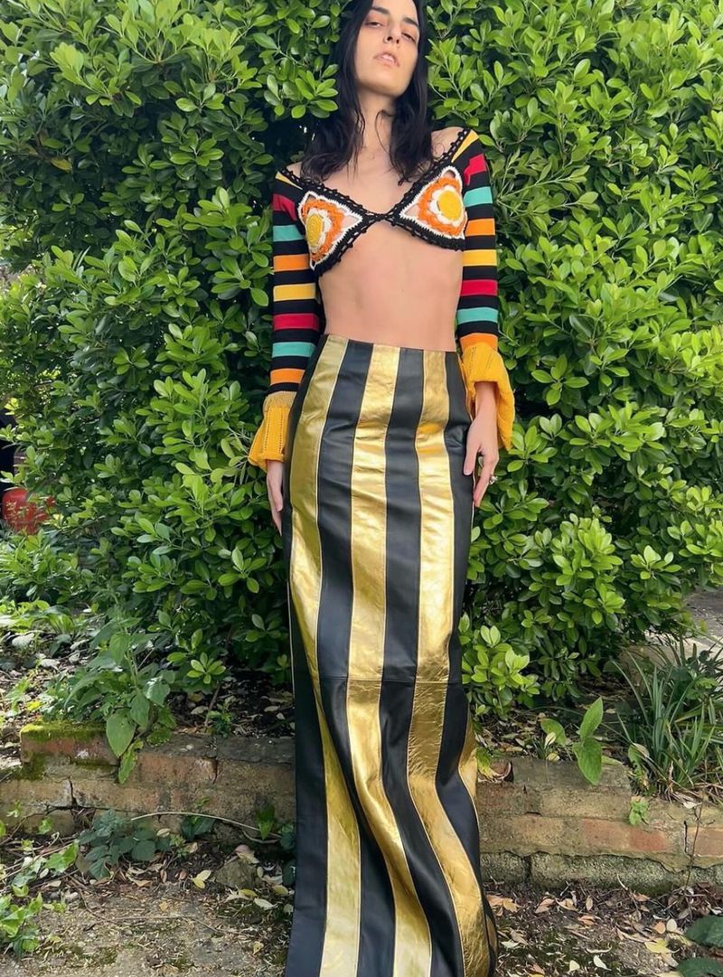 Adult woman wearing a colorful, crocheted crop top and a black and gold striped maxi skirt posing outdoors with a green leafy background.