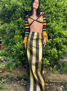 Model Nassia Matsa standing outdoors in a black and gold striped maxi skirt and multicolor knit crop top against lush green foliage.