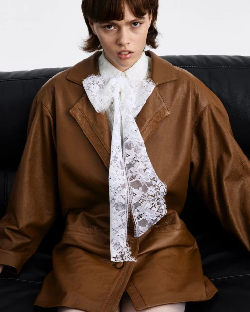 A young adult woman poses indoors wearing a brown leather-look suit and a white lace bow. The shot is a medium shot.