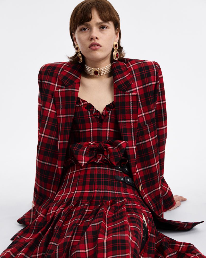 A young woman wearing a red and black plaid blazer and dress sits on a white surface, accessorized with statement earrings and a pearl choker.