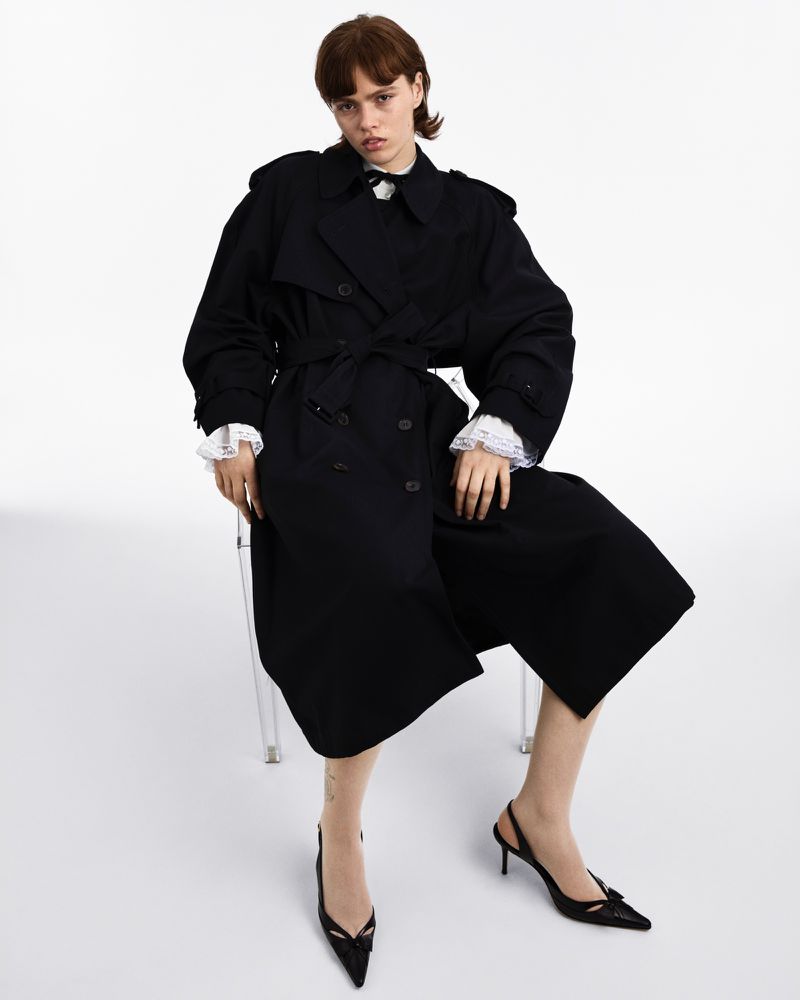 A young woman poses in a black trench coat and black heels in a studio setting. The image showcases fashion and style.