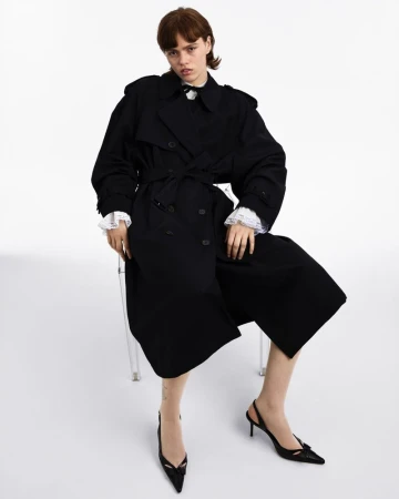 A young woman poses in a black trench coat and black heels in a studio setting. The image showcases fashion and style.