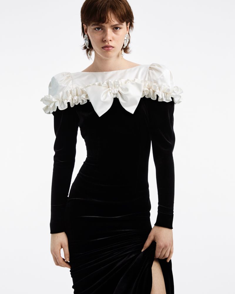 Woman in a long-sleeve black velvet dress with a white ruffled collar. The dress has a thigh-high slit. Studio shot.