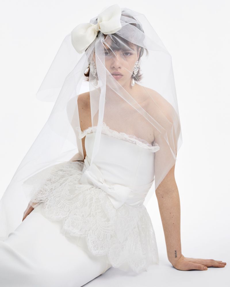 Woman wearing a white bridal dress with veil and a large decorative bow on her head, posing against a white background in a studio.