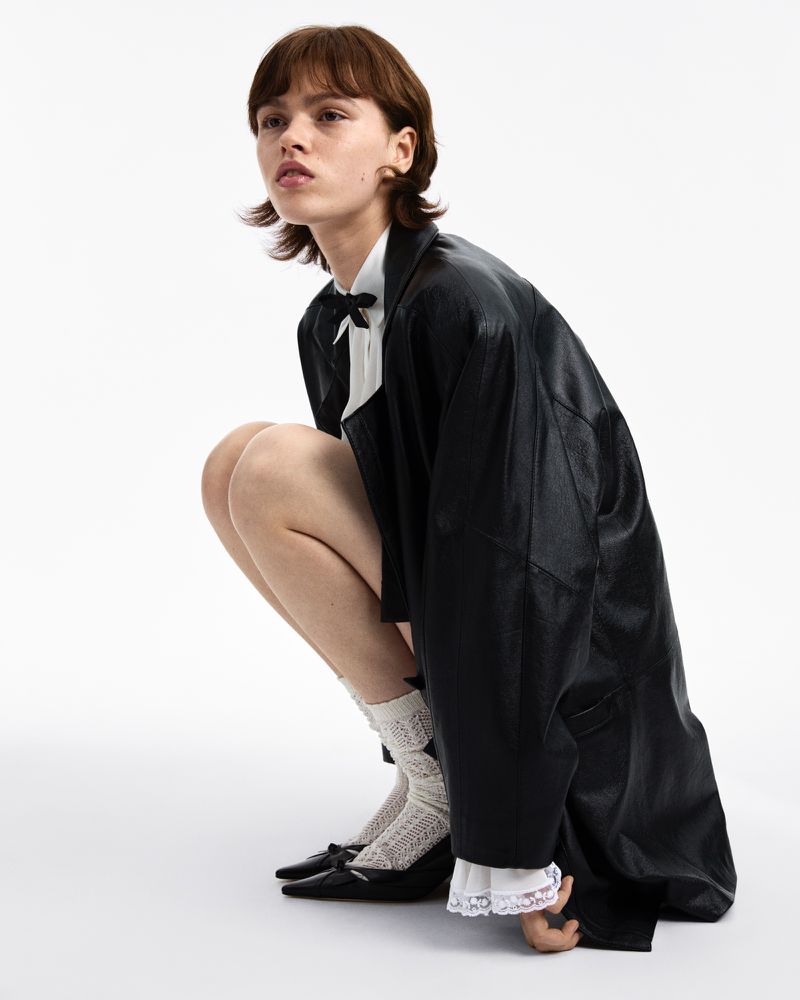 Woman in a black leather coat and white blouse poses in a studio, wearing lace socks and black heeled shoes against a white backdrop.
