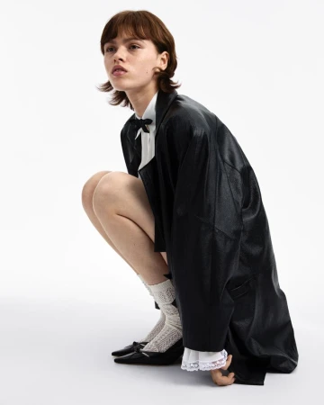 Woman in a black leather coat and white blouse poses in a studio, wearing lace socks and black heeled shoes against a white backdrop.