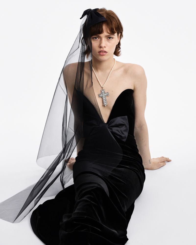 Elegant woman poses in a strapless black velvet gown with a bow detail, complemented by a pearl necklace and a delicate black veil headpiece.