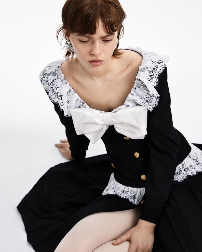 Woman poses seated wearing a black long-sleeve dress with a white lace collar and bow against a white backdrop in a studio setting.