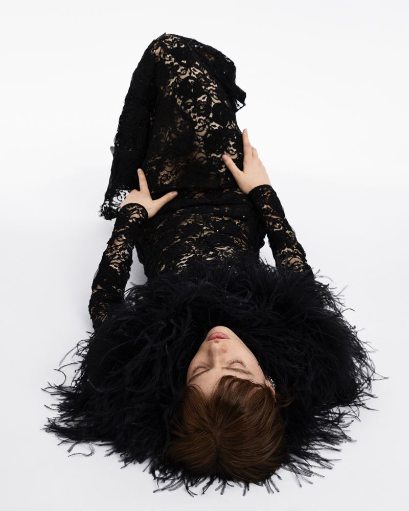 Woman in lace dress and feather boa lies on white background. Fashion portrait with elegant lace textures, soft lighting, and neutral colors.