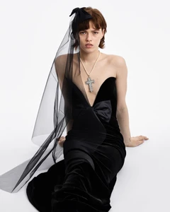 Model wearing a black velvet strapless gown and sheer veil with a crucifix necklace against a plain white background